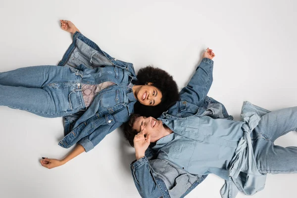 Vista superior de casal interracial feliz em roupas jeans deitado no fundo cinza — Fotografia de Stock