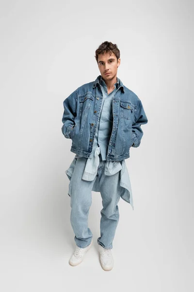 Полная длина стильного молодого человека в джинсовой куртке и белых кроссовках позирует с руками в карманах на сером — стоковое фото