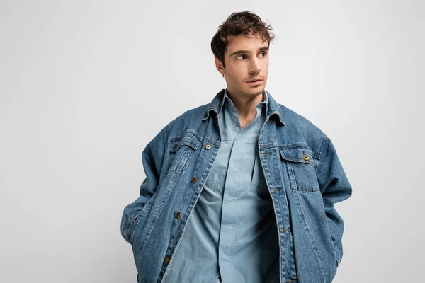 Stylish young man posing with hands in pockets of denim jacket isolated on grey — Stock Photo