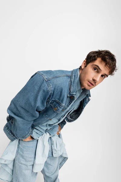 Stylish young man in denim clothes posing with hands in pockets and looking at camera isolated on grey - foto de stock
