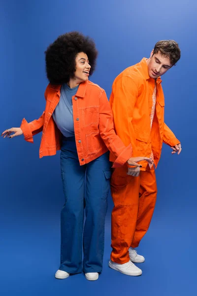 Усміхнена афроамериканка стоїть біля стильного чоловіка в помаранчевій куртці на синьому фоні — стокове фото