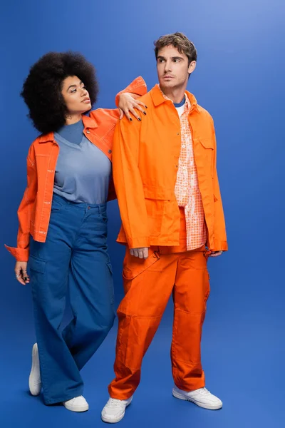 Full length of stylish multiethnic couple in jackets posing on blue background — Fotografia de Stock