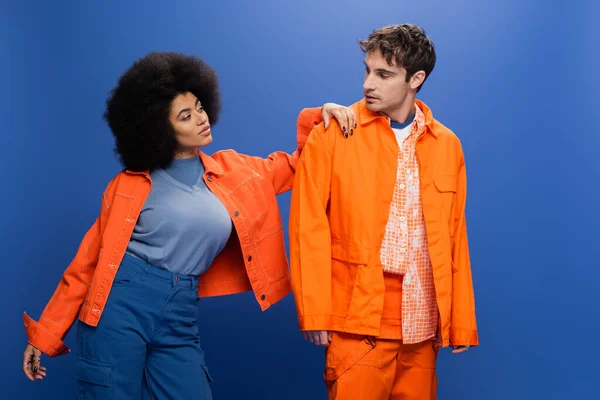 Trendy african american woman in bright jacket looking at man isolated on blue — Fotografia de Stock