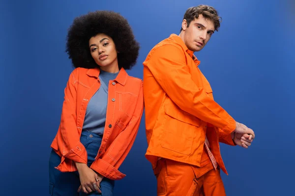 Модна багатоетнічна пара в помаранчевих куртках позує ізольовано на синьому — стокове фото