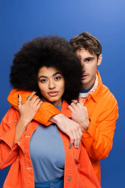 Чоловік в помаранчевій куртці обіймає афроамериканську жінку ізольовано на синьому — стокове фото