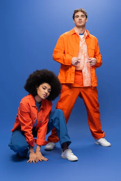 Впевнені міжрасові моделі в помаранчевих куртках, які дивляться на камеру на синьому фоні — стокове фото