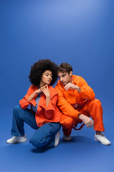 Молода багатоетнічна пара в помаранчевих куртках позує на синьому фоні — стокове фото