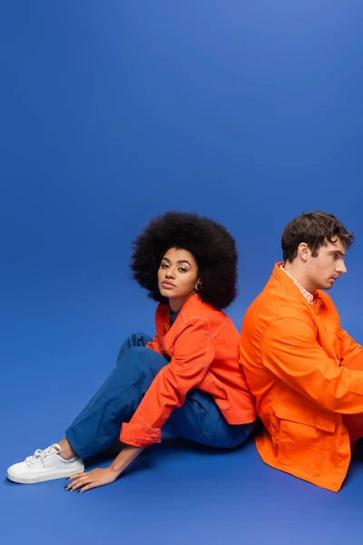 Curly african american woman in bright orange jacket sitting near man on blue background — Stockfoto