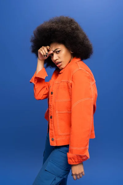 Trendy african american woman in orange jacket looking at camera isolated on blue — Stockfoto