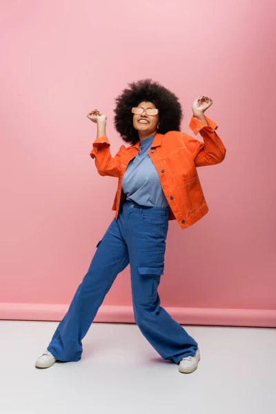 Cheerful african american woman in sunglasses and stylish outfit on pink background — Stock Photo