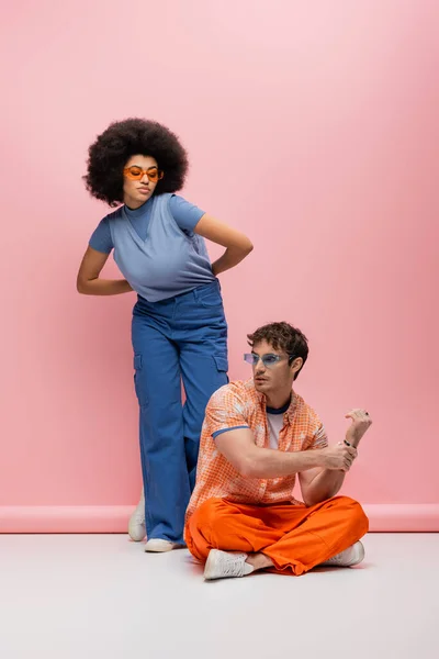 Stylish african american woman in blue clothes looking at brunette man in sunglasses on pink background — Stockfoto