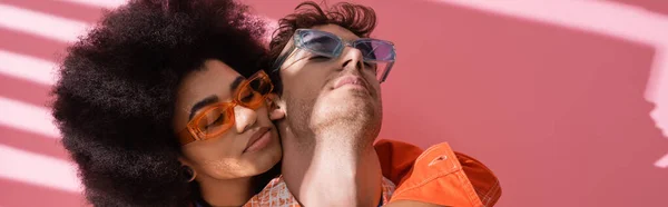 Young african american woman hugging man in sunglasses on pink background with shadow, banner — Stock Photo