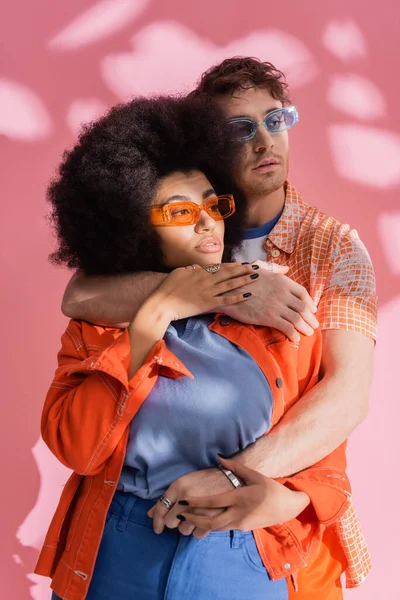 Stylish brunette man hugging african american woman on pink background with shadow — Stockfoto