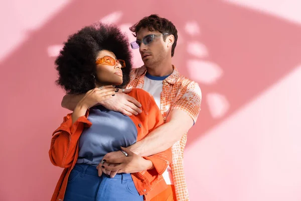 Man in sunglasses hugging stylish african american woman on pink background with shadow — Foto stock