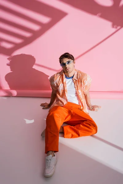 Modelo con estilo en general y gafas de sol sentadas sobre fondo rosa con sombra - foto de stock
