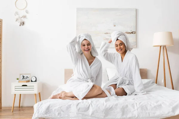 Fröhliche multiethnische Frauen in weißen Bademänteln und Handtüchern, die auf dem Bett in die Kamera schauen — Stockfoto