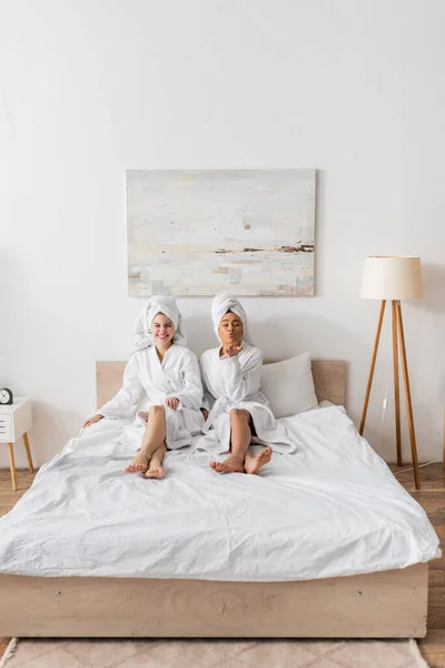 Full length of african american woman blowing air kiss near cheerful friend in bathrobe and towel on bed at home — Stock Photo