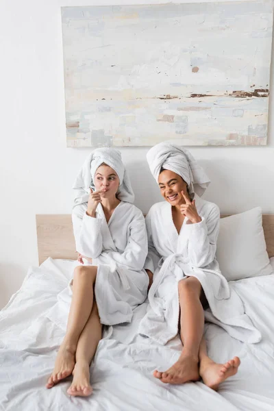 Full length of cheerful interracial women in white bathrobes and towels smiling and grimacing on bed at home - foto de stock