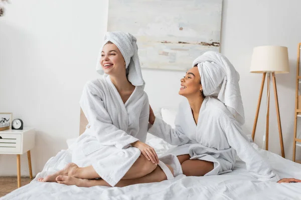 Happy young woman in terry bathrobe and towel looking away near smiling african american friend on bed - foto de stock