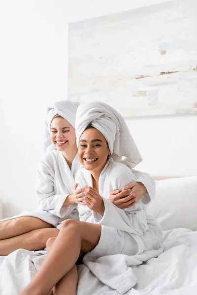 Cheerful african american woman in white bathrobe and towel looking at camera near happy friend in bedroom - foto de stock