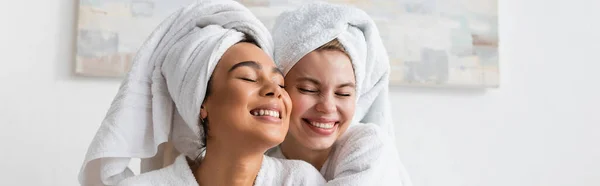 Pretty and cheerful interracial women in white robes and towels smiling with closed eyes in bedroom, banner - foto de stock