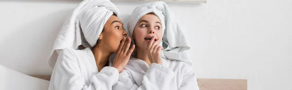 African american woman in terry robe and towel telling gossip to surprised friend in bedroom, banner — Stockfoto