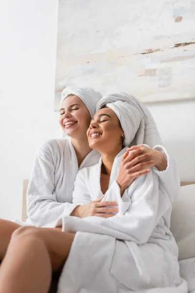 Junge und glückliche interrassische Frauen in weißen Frottee-Roben und Handtüchern, die sich mit geschlossenen Augen im Schlafzimmer umarmen — Stockfoto
