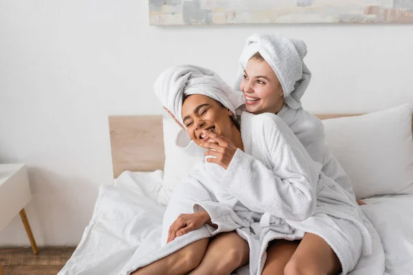 Excited african american woman in white bathrobe and towel laughing with closed eyes near happy friend on bed - foto de stock