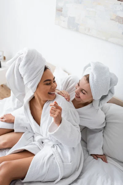 High angle view of young multiethnic women in white towels and bathrobes smiling at each other on bed - foto de stock