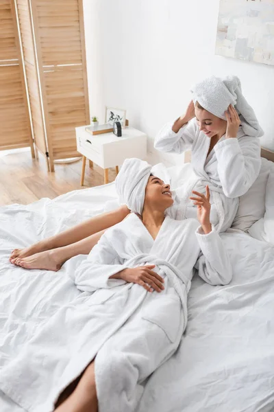 Mujer afroamericana en bata blanca hablando con un amigo sonriente ajustando la toalla en la cabeza en el dormitorio - foto de stock