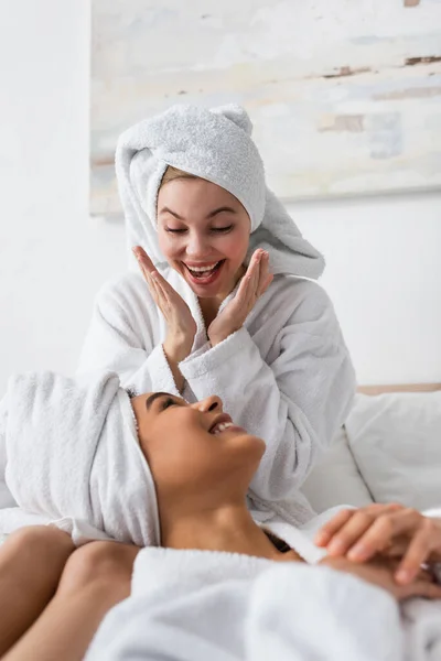 Excited woman in white soft bathrobe and towel showing wow gesture near smiling african american friend in bedroom — Photo de stock