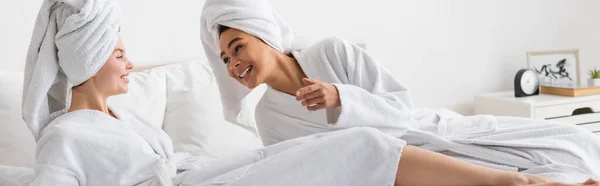 Cheerful african american woman in white soft bathrobe and towel talking to young friend in bedroom, banner - foto de stock
