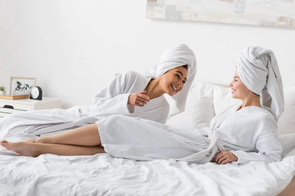 Cheerful african american woman talking to pretty friend lying on bed in white terry bathrobe and towel - foto de stock