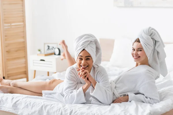Fröhliche multiethnische Frauen entspannen sich in weißen Frottee-Bademänteln und Handtüchern, während sie in die Kamera schauen — Stockfoto