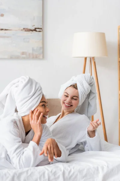 Joyful interracial women in white bathrobes and towels gesturing during conversation in bedroom - foto de stock