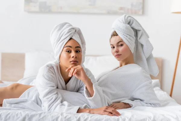 Junge und hübsche interrassische Frauen in weißen Frottee-Bademänteln und Handtüchern, die zu Hause auf dem Bett in die Kamera schauen — Stockfoto