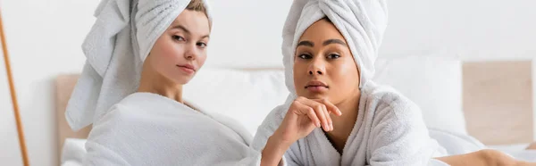Young multiethnic women in white bathrobes and towels looking at camera in bedroom, banner - foto de stock