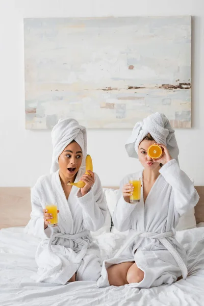 Young woman pouting lips and covering eye with ripe orange near african american woman with banana and orange juice — Foto stock