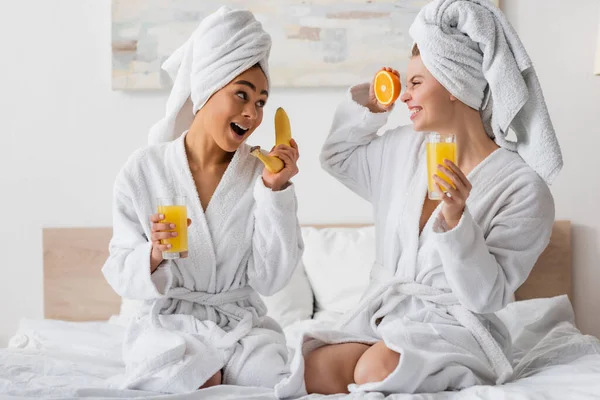Excited multiethnic women with fresh fruits and orange juice having fun while sitting on bed — Foto stock