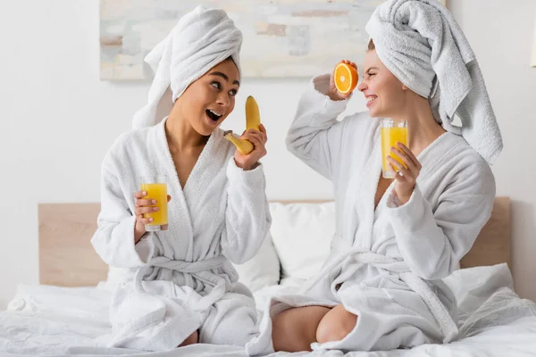 Eccitate donne multietniche con frutta fresca e succo d'arancia divertirsi mentre si siede sul letto — Foto stock