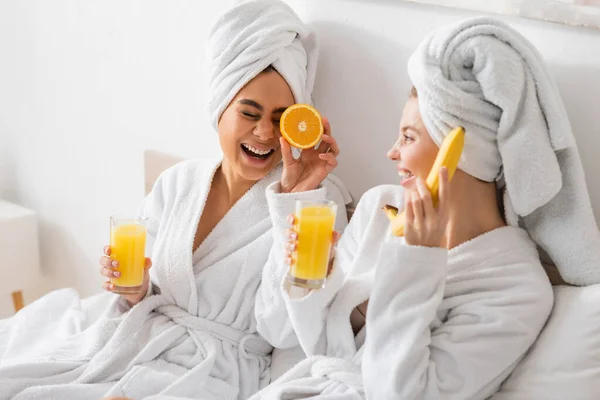Young multiethnic women in white robes and towels having fun with ripe fruits in bedroom — Foto stock
