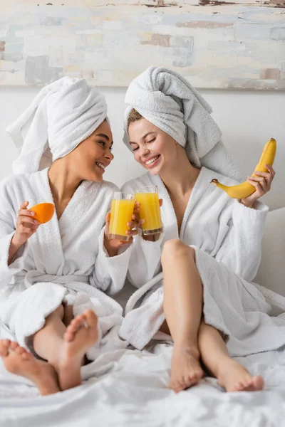 Full length of barefoot interracial women in white robes clinking glasses of orange juice while sitting on bed — Photo de stock