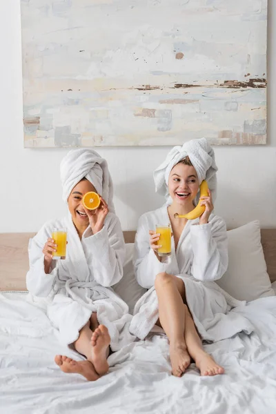 Longitud completa de alegres mujeres interracial con frutas y jugo de naranja divertirse mientras se sienta en la cama - foto de stock