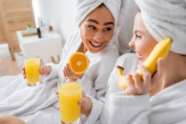 Happy african american woman holding ripe orange and juice near blurred friend having fun in bedroom — Foto stock