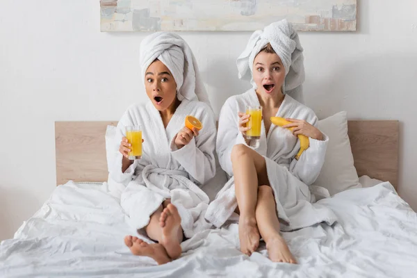 Full length of amazed interracial women in white robes and towels holding fruits and orange juice while looking at camera on bed — Foto stock