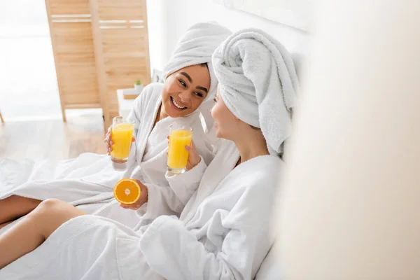 Joyful african american woman with ripe orange and juice looking at friend in bedroom on blurred foreground — Photo de stock