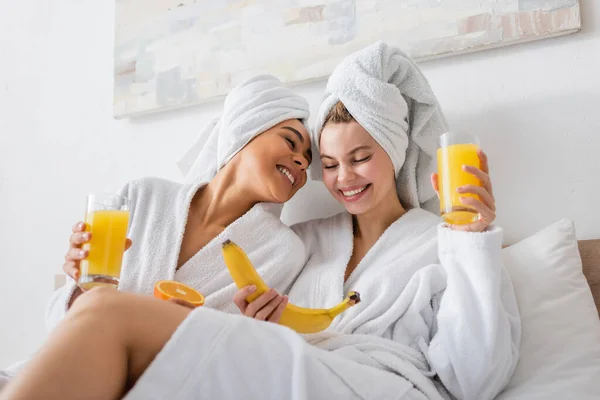 Young and happy multiethnic women in white bathrobes and towels holding fruits and fresh orange juice in bedroom — Foto stock