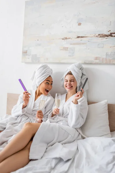 Mujer afroamericana asombrada con alegre amigo sosteniendo champán y limas de uñas mientras está sentado en batas blancas y toallas en la cama - foto de stock