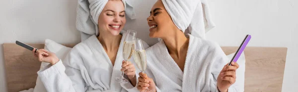 Femmes multiethniques heureux en éponge peignoirs et serviettes tenant des limes à ongles et des verres à champagne dans la chambre à coucher, bannière — Photo de stock