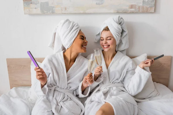 Alegres mujeres multiétnicas en batas y toallas de rizo blanco sosteniendo limas de uñas y tintineo copas de champán en la cama - foto de stock